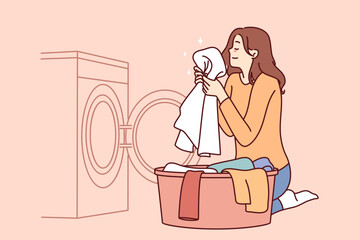 Housewife woman sits near washing machine and inhales fragrant smell freshly washed towel after using good laundry detergent. Girl takes out washed clothes from washing machine putting them in bowl 