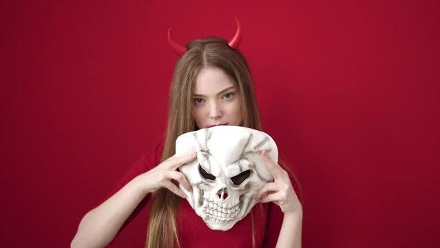 Young blonde woman wearing devil costume holding skull mask over isolated red background