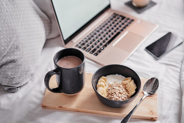 Breakfast in bed: a wooden tray with granola and cup of cocoa with a laptop on background.