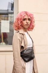Portrait shooting of a stylish girl with pink hair. Beige shades. Trends of spring and summer 2023.Beige Trench cloak with white top and pants. Black small belt bag