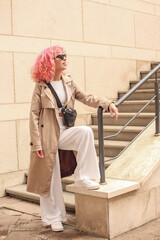 Portrait shooting of a stylish girl with pink hair. Beige shades. Trends of spring and summer 2023.Beige Trench cloak with white top and pants. Black small belt bag