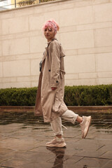 Portrait shooting of a stylish girl with pink hair. Beige shades. Trends of spring and summer 2023. Beige trench cloak with white t-shirt and beige pants. Black small belt bag