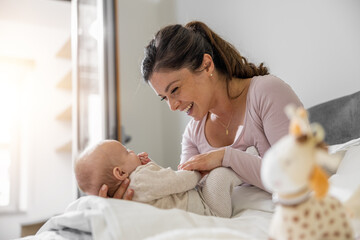 Portrait of beautiful mom playing with her three months old baby in bedroom - obrazy, fototapety, plakaty