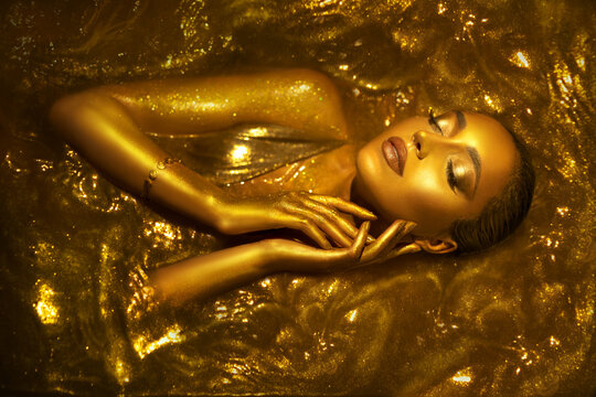 Portrait Closeup Beauty fantasy woman face in gold paint. Golden shiny skin. Glamorous fashion model girl image goddess lies bathed in liquid gold. Professional metallic glowing luxury royal makeup 