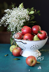 Red apples on black background. Vibrant colors of fresh seasonal fruit. Apples in a colander. White flowers and fruit still life. 