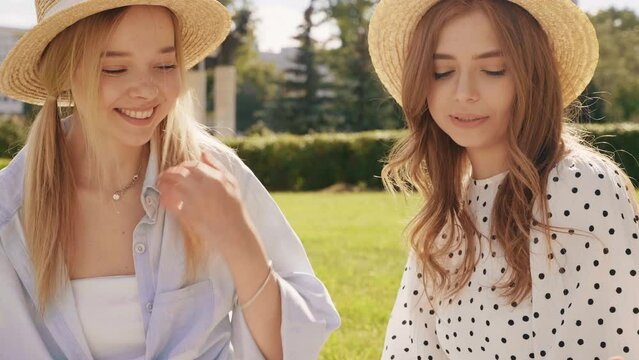Two young beautiful hipster woman in trendy summer sundress and hats. Carefree women making picnic outside. Positive models sitting on plaid on grass, eating fruits and cheese