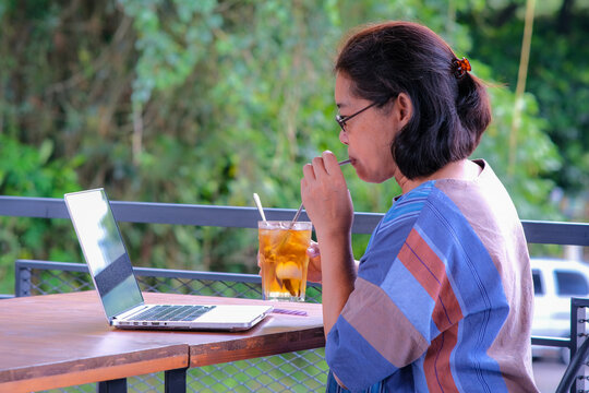 Asian woman drinking iced lemon tea while working on her laptop in her home terrace