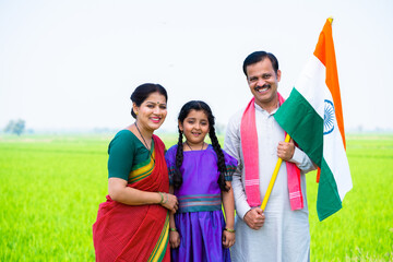 Happy smiling village couple with kid standing by holding indian flag by looking camera at farmland - concept of freedom, patriotism and family bonding