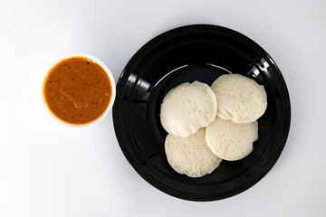 Hot and steaming idly in a black plate and tomato chutney  in a isolated white background. Steaming hot idly and red chutny isolated. Healthy food / Break fast in india.