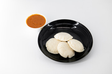 Hot and steaming idly in a black plate and tomato chutney  in a isolated white background. Steaming hot idly and red chutny isolated. Healthy food / Break fast in india.