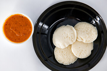 Hot and steaming idly in a black plate and tomato chutney  in a isolated white background. Steaming hot idly and red chutny isolated. Healthy food / Break fast in india.