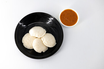 Hot and steaming idly in a black plate and tomato chutney  in a isolated white background. Steaming hot idly and red chutny isolated. Healthy food / Break fast in india.