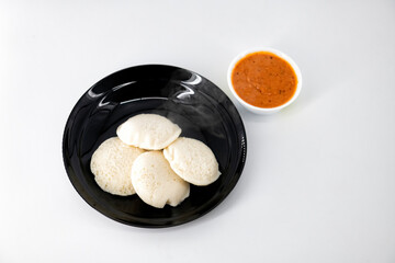 Hot and steaming idly in a black plate and tomato chutney  in a isolated white background. Steaming hot idly and red chutny isolated. Healthy food / Break fast in india...