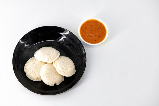 Hot and steaming idly in a black plate and tomato chutney  in a isolated white background. Steaming hot idly and red chutny isolated. Healthy food / Break fast in india.