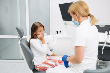 Fototapeta premium Little girl suffering from toothache and sitting at dental chair before teeth examining by dentist.