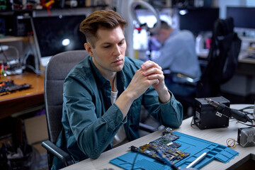 confident young IT specialist have rest after repair at workshop, hardware upgrading. caucasian serious guy sit behind table with disassembled equipment, looking away thinking