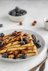 Crepes or blinis or thin pancakes with blueberries, hazelnuts and chocolate sauce on a white plate at white stone table. Maslenitsa traditional festival meal.