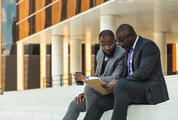 Friendly meeting of business partners outdoors. Two dark-skinned men in suits sit on the steps of a city building with a notebook and have a conversation. Working break. Support and collaboration