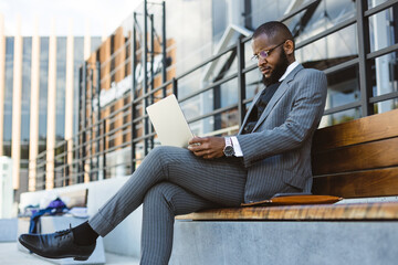 A dark-skinned man in a business suit is using a laptop outdoors. Digital education and remote work
