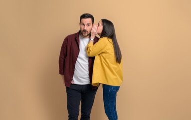 Side view of girlfriend hiding her mouth and telling secret to surprised boyfriend. Young couple sharing gossips with each other while standing isolated on beige background