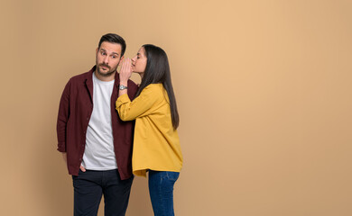 Side view of young woman hiding her mouth while telling secret to her charming boyfriend. Couple sharing gossips with each other while standing together on isolated beige background