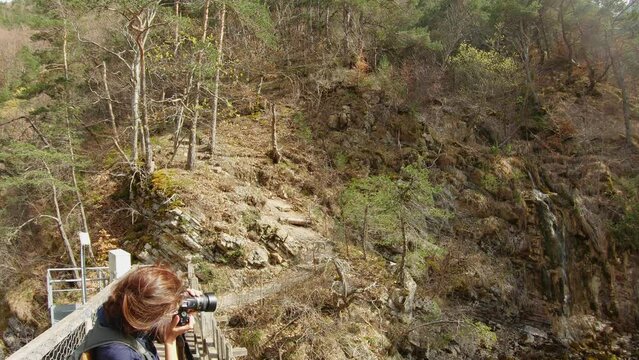 young caucasian woman on a bridge taking picture of a small waterfall. She uses a DSLR camera. Video in slow motion