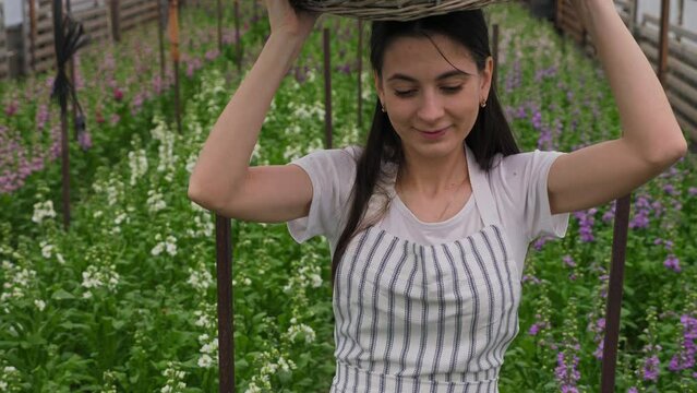 The girl carries a wicker basket with matthiola flowers on her head, looking down and smiling.