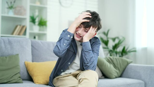 Teen child with tension, boy suffering from headache sitting on couch at home indoor upset exhausted teenager guy feeling frustrated depressed tired touching forehead suffer with anxiety mental health
