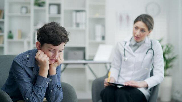Pediatrician doctor comforting a sad boy in the office of a hospital or clinic. Woman physician having conversation to soothe with sorrowful, dismal depressed patient Female therapist consoling child