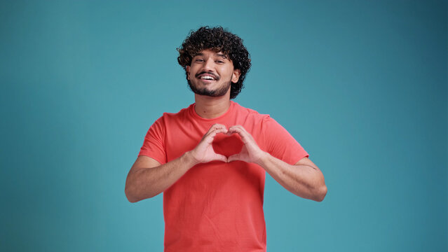 Handsome curly millennial hindu guy wearing coral t-shirt showing heart-shaped gesture over his chest and smiling at camera isolated on a blue studio background, copy space. Love, emotions concept
