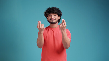 Naklejka premium Young handsome Indian man with beard in coral t-shirt on blue studio background doing money gesture with hands, asking for salary payment