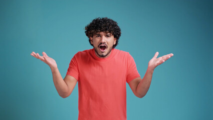 Shocked young bearded Indian man 20s years old says what. wears coral t-shirt on blue studio...