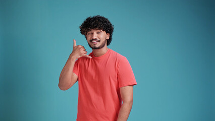 Young Indian man wearing in coral t-shirt on blue studio background smiling at camera and shows call me telephone hand sign gesture