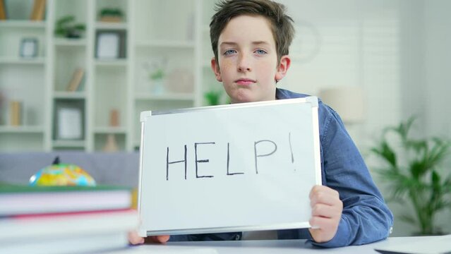 Portrait of a tired schoolboy who sits and teaches lessons at the table at home. Showing a board that says Help A teenage boy exhausted by schoolwork task boring Looks into the camera
