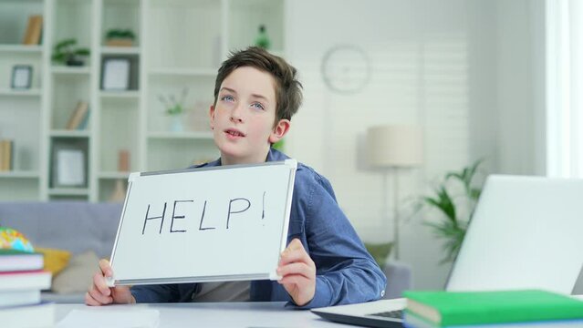 Portrait of a tired schoolboy who sits and teaches lessons at the table at home. Showing a board that says Help A teenage boy exhausted by schoolwork task boring Looks into the camera