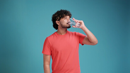 Indian man drinks water from a glass in coral t-shirt on blue studio background