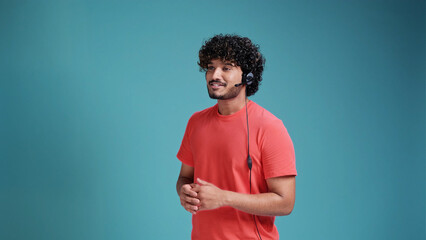 indian male call center operator with head set talking in coral t-shirt on blue studio background