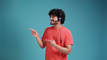 Young indian call center agent man using headset in coral t-shirt on blue studio backgroun pointing and showing with thumb up to the side with happy face smiling