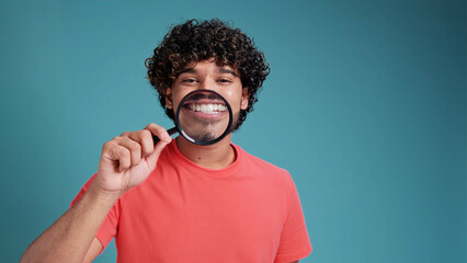 funny indian man shows white teeth and smiles through a magnifying glass in coral t-shirt on blue studio background