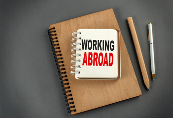 WORKING ABROAD text on notebook with pen and pencil on grey background