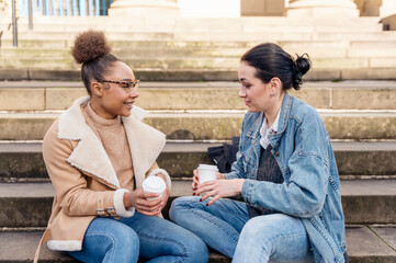 two student women in a denim jacket is talking to each other, on phone, laughing, drinking coffee  on stairs in city.