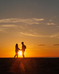Fototapeta premium silhouette of a couple at sunset, woman and man walking along seashore in rays of sunset sunlight. walk and hug. place for text, romantic relationships, freedom and carelessness