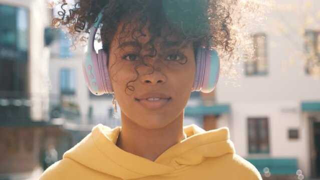 Beautiful black woman with afro curls hairstyle. Smiling model in yellow hoodie. Sexy carefree female enjoying listening music in wireless headphones. Posing on the street background at sunset