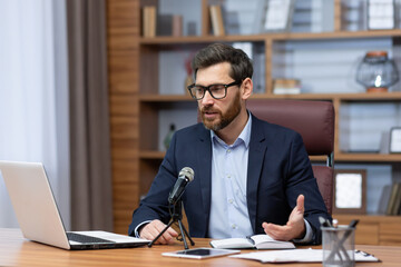 Mature businessman inside office with professional microphone and laptop recording audio podcast and training seminar, experienced boss financier sitting at desk in office video call online stream.