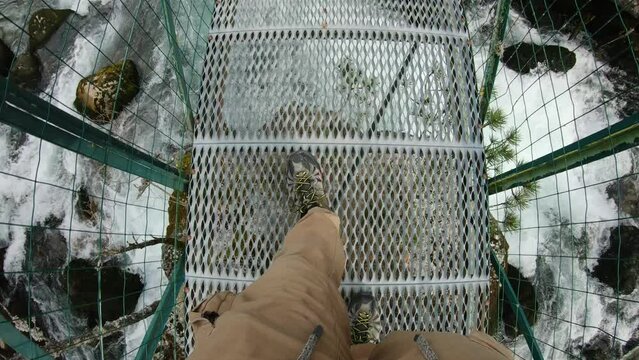 First person view looking down at is feet while crossing a metal bridge over a powerfull river. Slowmotion