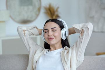 Young beautiful woman in headphones sitting on the sofa at home with closed eyes and hands behind her head and resting, listening to music.