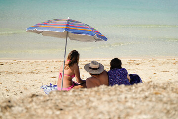 Unrecognizable backward tourist family in the beach with a parasol in the sand in summertime...