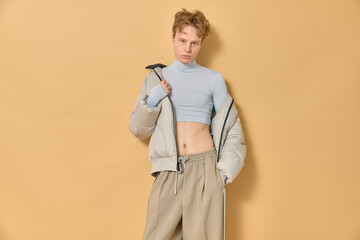 Queer man with red curly hair wearing grey long sleeved cropped turtleneck beige pants and winter...