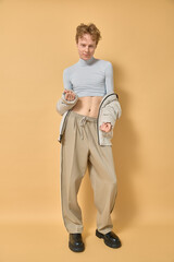 Modern man vogue hip-hop dancer in motion wearing stylish clothing beige pants crop top and coat showing dance movements standing with bare belly smiling fooling around over yellow background. 
