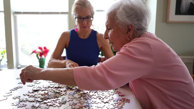 Happy pretty elderly woman moves puzzle pieces while daughter organizes pieces to fit together. Concept of spending time with loved ones.